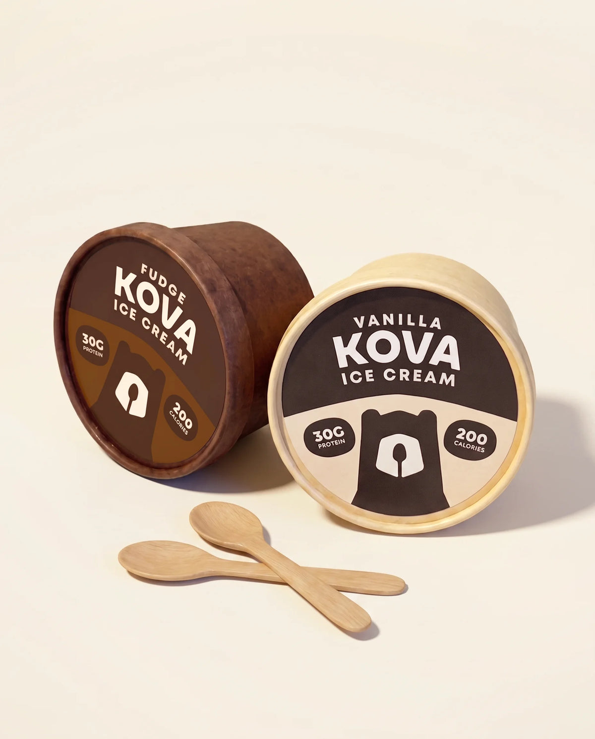 Kova Fudge and Vanilla protein ice cream cups with wooden spoons on a cream background