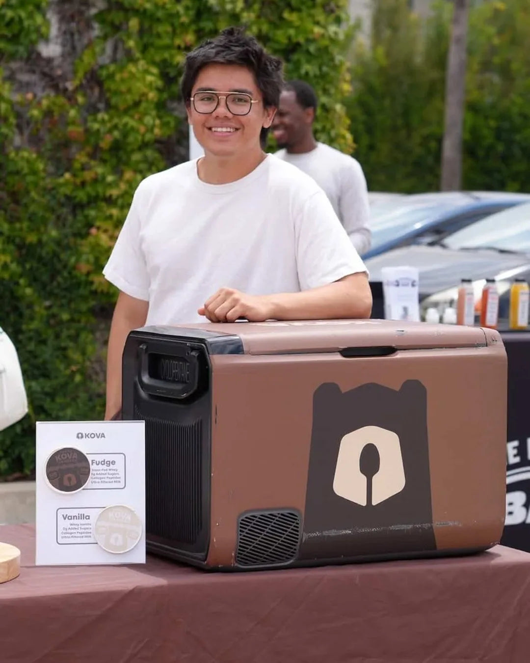 Kova founder Juan Turcios smiling behind the brown Kova-branded freezer at an outdoor pop-up table, with a Fudge and Vanilla flavour card beside it
