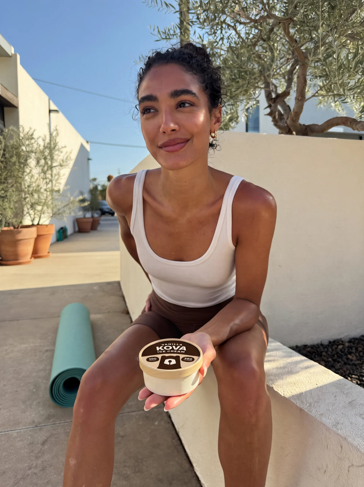 A woman resting on a sunny wall after a workout, holding a cup of Kova Vanilla protein ice cream