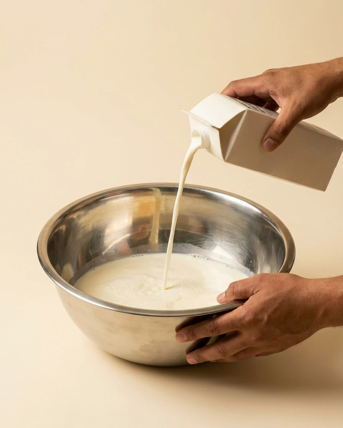 Hands pouring milk from a carton into a steel mixing bowl — the start of a batch of Kova